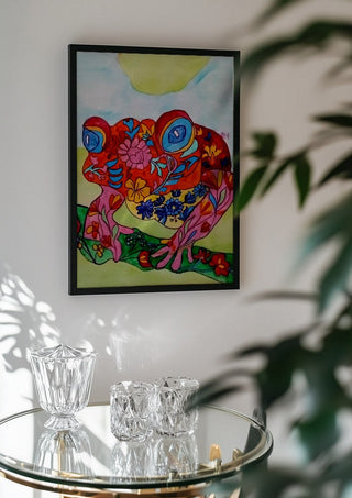 Color frog by Nittaart in a black frame above a glass table with crystal glassware