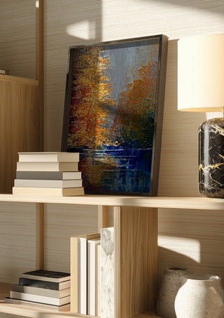 Autumn on the Water by Justyna Kopania in a dark frame on a wooden shelf beside books and a lamp
