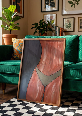 Bare Confidence by Bibi in a wooden frame leaning against a green velvet sofa on a chequered floor