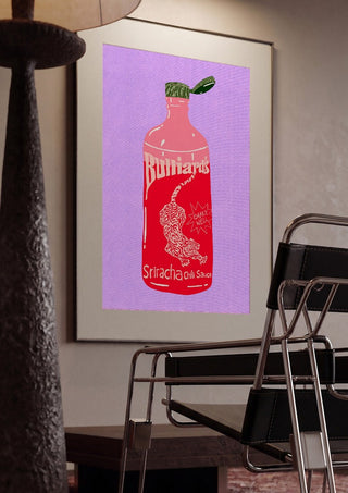 Sriracha chili sauce by Astrid Lind framed in a dim room beside a tubular steel chair