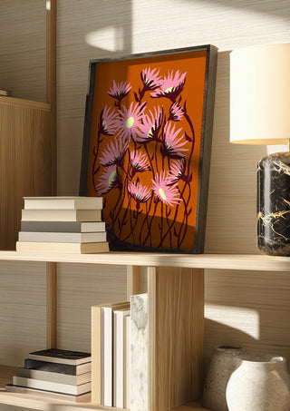 Whimsical flowers by Astrid Lind in a dark frame on a wooden shelf beside books and a lamp