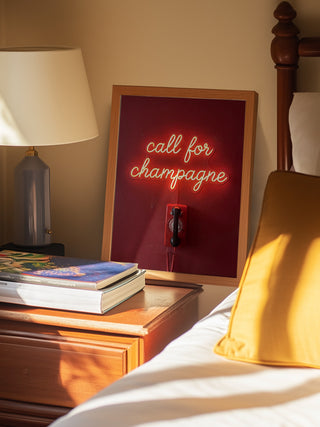 Neon sign reading "call for champagne" by Andreas Magnusson on a nightstand next to a bed.