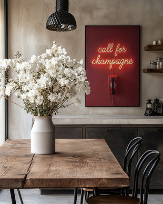 A "Call For Champagne" neon sign by Andreas Magnusson on a wall above a dining table with white flowers.