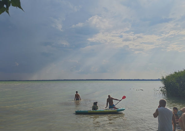 The Lake’s Shared Rhythm by Bora Kőhalmi - swimmers and a paddleboarder on the green water of Lake Balaton under a wide cloudy sky