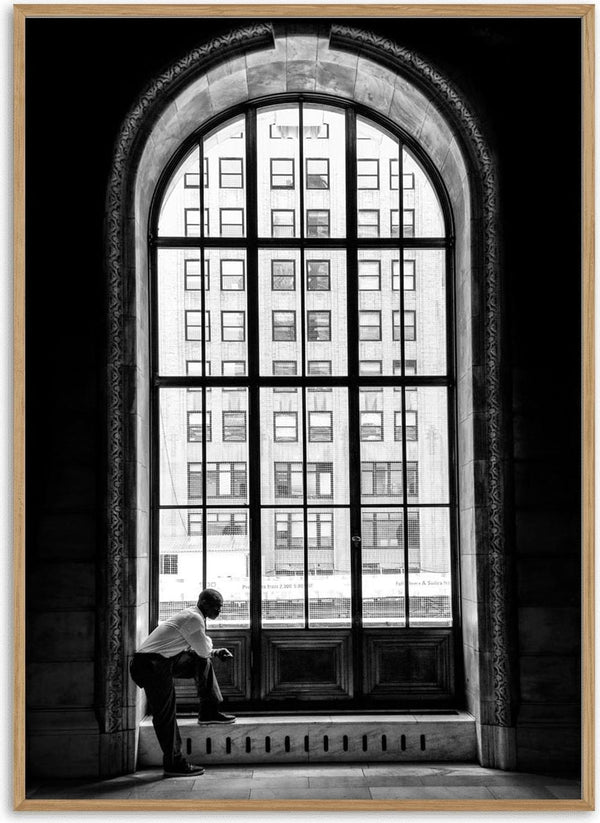 A man stands alone by a tall arched window, gazing at city buildings outside.